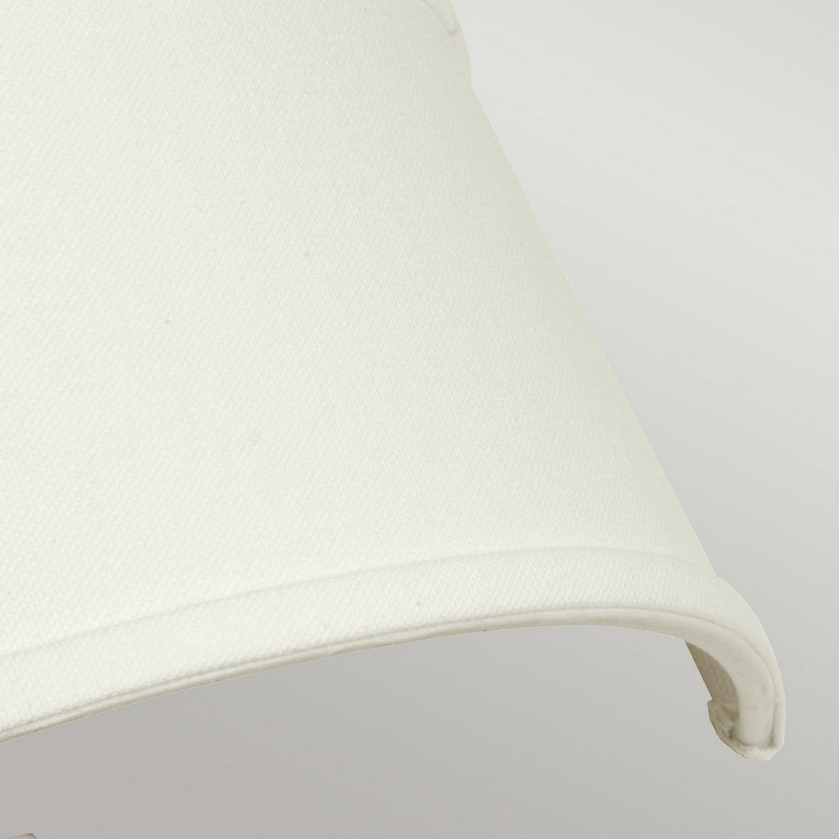 Close-up of an ivory linen drum shade on a light grey background. The fabric texture is visible, highlighting the smooth, rounded edge of the shade. This elegant detail beautifully complements the Antique Nickel finish of the Hampton 2 Light Wall Light - Antique Nickel With Ivory Shade.