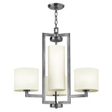 The Hampton 4 Light Chandelier - Antique Nickel with Ivory Shade showcases a contemporary, transitional design featuring three cylindrical ivory lampshades. These shades are arranged in a triangular formation and suspended by a chain from a rectangular frame. The central shade is larger, contributing an elegant touch to the fixture's overall appearance.