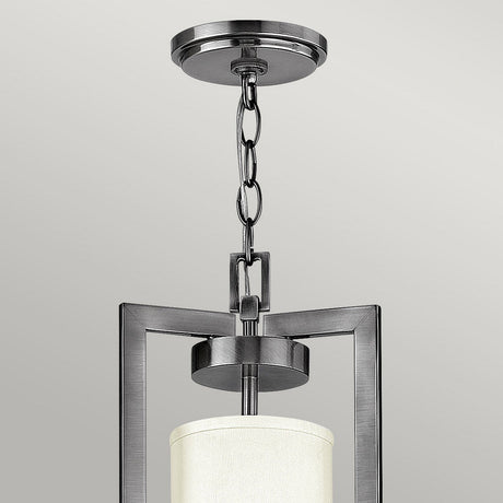 The Hampton 4 Light Chandelier, featuring an Antique Nickel finish and an Ivory shade, showcases a modern pendant design with a brushed metal frame and cylindrical lampshade. Its sleek look is reminiscent of Transitional Design, as it hangs elegantly from a chain attached to a ceiling mount.