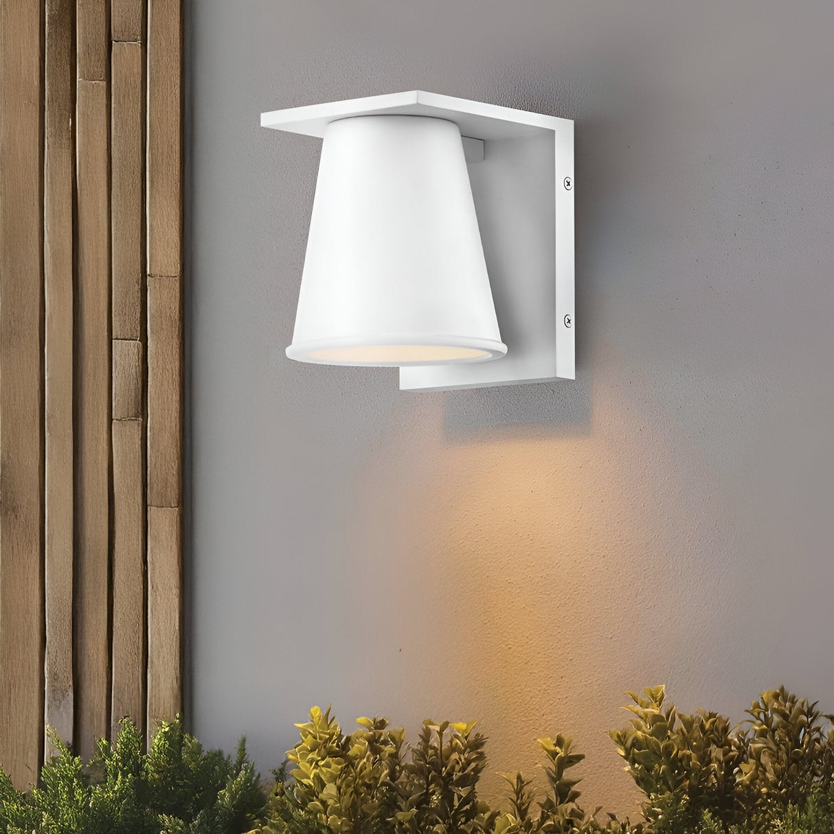The Coastal 1 Light Small Outdoor Wall Lantern in textured white, featuring a conical shade, is mounted on a gray wall and shines light downward onto green plants. Vertical wooden slats are visible on the left side of the image.