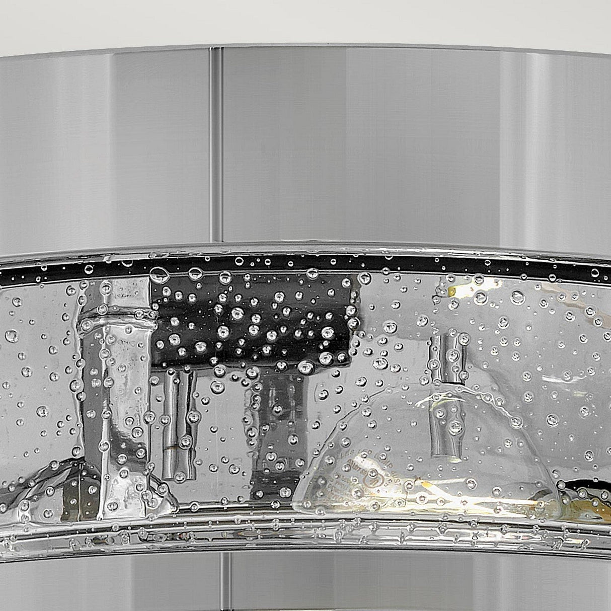 Close-up of the Harper 3 Light Flush in polished chrome with clear seeded glass. Water droplets create a pattern across its curved, reflective surface, adding texture and depth. The sleek metallic finish exudes modernity, enhanced by a subtle sheen.