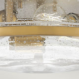 A close-up of the Harper 3 Light Flush fixture, featuring a Heritage Brass frame and clear seeded glass, adorned with droplets of water. The blurred interior background enhances its retro design charm with reflections and shadows.