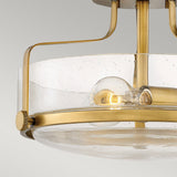 The Harper 3 Light Small Semi Flush in Heritage Brass with Clear Seeded Glass showcases a contemporary design, featuring clear seeded glass and heritage brass accents. This semi-flush mount fixture reveals a visible incandescent bulb, while its brass arms elegantly curve to support the design against a plain light gray background.