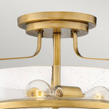 Close-up of the Harper 3 Light Small Semi Flush, showcasing its elegant design with a brushed Heritage Brass finish. It features an open circular clear seeded glass shade that encloses three exposed bulbs, suspended from a circular base with curved supports in a stylish semi flush mount design.