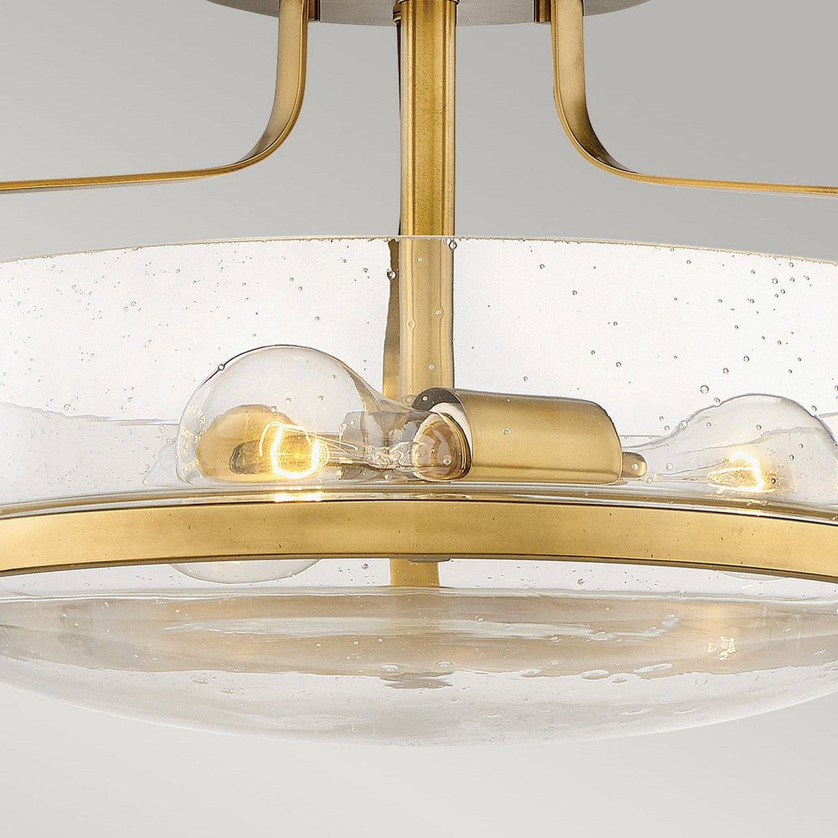 Close-up of the Harper 3 Light Small Semi Flush in Heritage Brass with a circular, clear seeded glass shade. The fixture features two exposed light bulbs that emit a warm glow against a neutral background.