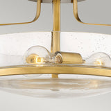 Close-up of the Harper 3 Light Small Semi Flush in Heritage Brass with a circular, clear seeded glass shade. The fixture features two exposed light bulbs that emit a warm glow against a neutral background.