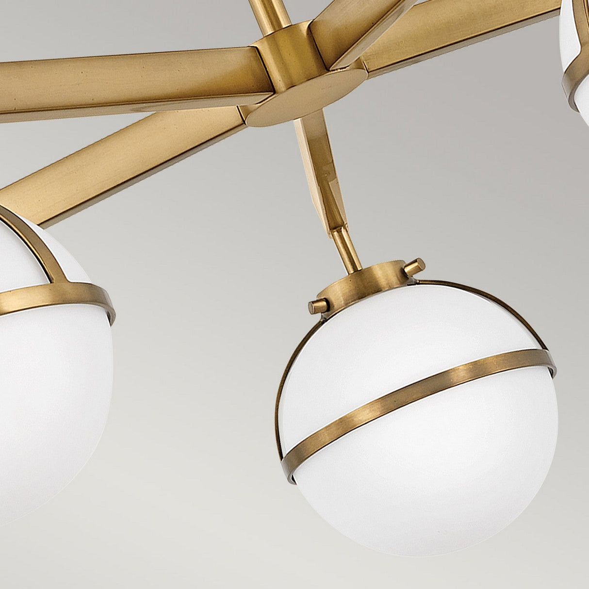 Close-up of the Hollis 5 Light Semi Flush chandelier in Heritage Brass, showcasing a modern brushed brass finish. The design features opal etched glass shades encircled by brass bands and connected by matching metal rods, set against a soft gray background.