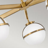 Close-up of the Hollis 5 Light Semi Flush chandelier in Heritage Brass, showcasing a modern brushed brass finish. The design features opal etched glass shades encircled by brass bands and connected by matching metal rods, set against a soft gray background.