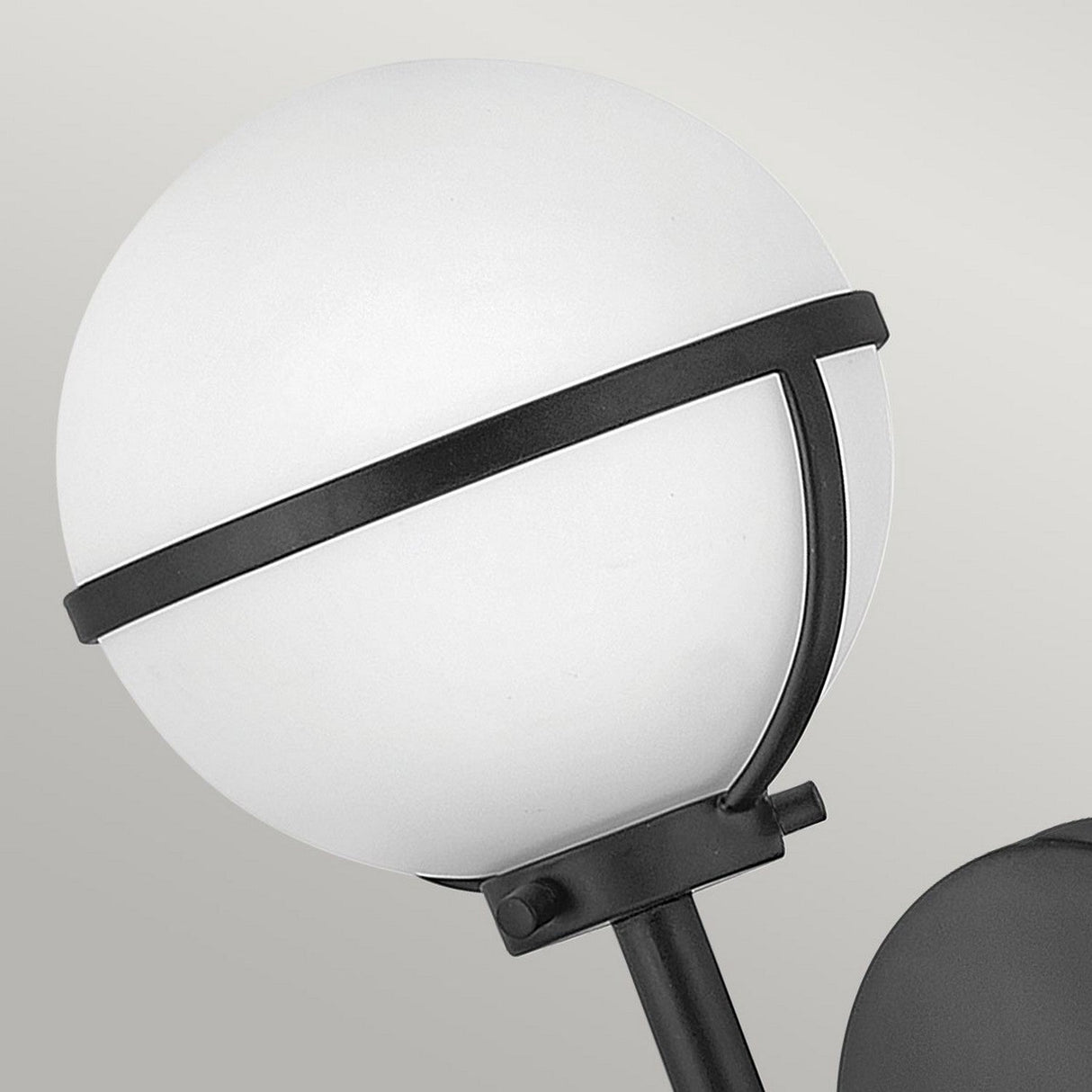 The Hollis 1 Light Wall Light, designed with Art Deco inspiration, showcases a spherical opal glass globe encircled by a black metal band and is mounted on a sleek black stand against a light gray background.