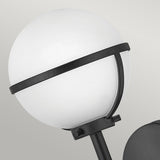 The Hollis 1 Light Wall Light, designed with Art Deco inspiration, showcases a spherical opal glass globe encircled by a black metal band and is mounted on a sleek black stand against a light gray background.