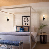 A stylish Art Deco bedroom showcases a white canopy bed decorated with blue and patterned pillows. A framed abstract portrait is displayed on a textured wall, illuminated by the soft light of the Hollis 1 Light Wall Light in black with opal glass. Beside the bed sits a small round table topped with a vase of leaves.