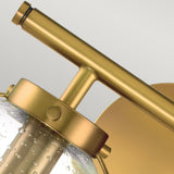 A detailed view of the Hollis 2 Light Wall Light in Heritage Brass showcases its cylindrical clear glass shade, which contains small air bubbles for added texture. This elegantly designed fixture features a sleek horizontal arm and a circular wall mount.
