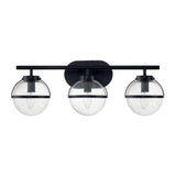 The Hollis 3 Light Wall Light in black with clear glass showcases a contemporary Art Deco style. It features three globe lampshades made of clear glass, mounted on a horizontal black bar. Each globe encases a filament bulb that highlights its sleek and elegant design.
