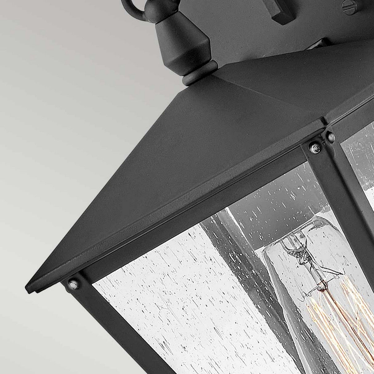Close-up of the Huntersfield Outdoor 1 Light Small Wall Lantern in a black finish, featuring clear glass panels. This outdoor lighting piece displays a visible light bulb inside and is mounted on a light-colored wall, with raindrops on the glass suggesting recent or ongoing rain.