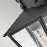Close-up of the Huntersfield Outdoor 1 Light Small Wall Lantern in a black finish, featuring clear glass panels. This outdoor lighting piece displays a visible light bulb inside and is mounted on a light-colored wall, with raindrops on the glass suggesting recent or ongoing rain.