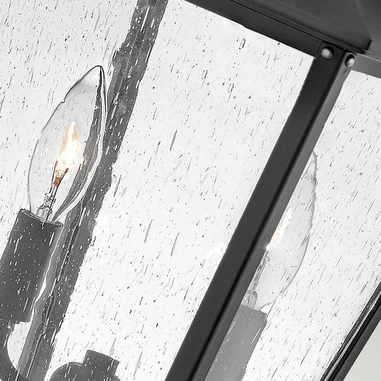 Close-up of the Huntersfield Outdoor 2 Light Medium Pedestal Lantern in Black, showcasing a lit decorative candle-style bulb within a glass enclosure framed by metal. The speckled water droplets on the glass hint at its IP44 rating, indicating its design to withstand rainy days, making it an ideal choice for an outdoor lantern.