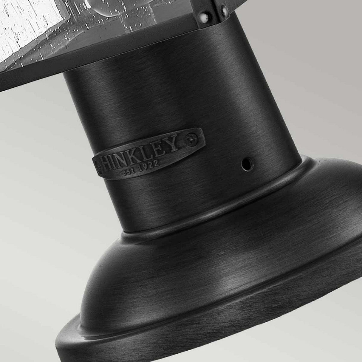 Close-up of the Huntersfield Outdoor 2 Light Medium Pedestal Lantern in black, showcasing HINKLEY and EST. 1922 embossed on a metal plate. This modern IP44 rated fixture features a textured, cylindrical base against a soft gray background, effortlessly combining style and practicality.