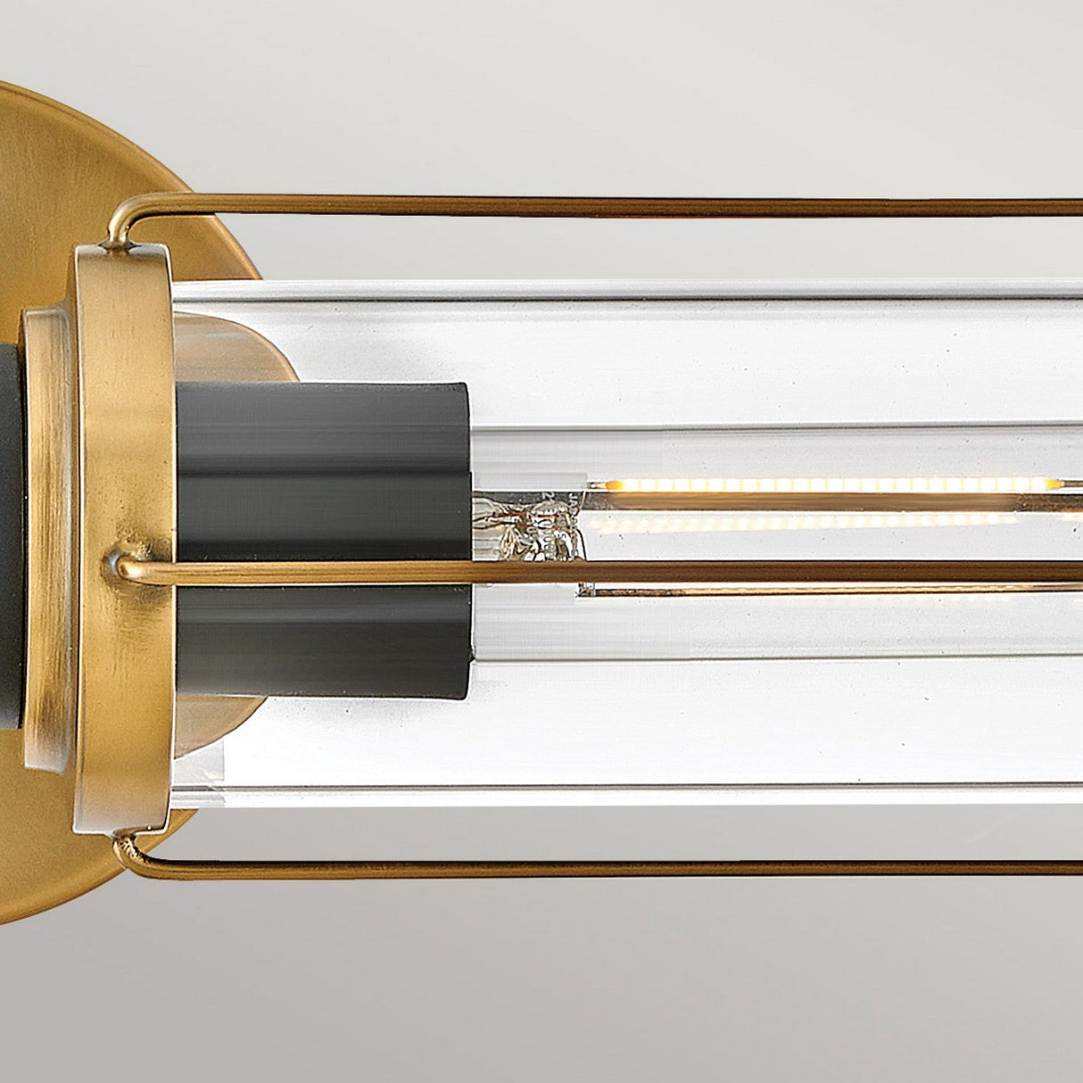The Masthead 2 Light Wall Light, featuring a cylindrical clear glass shade and exposed LED filament, is showcased in detail. It's adorned with Heritage Brass finish accents alongside black elements, combining sleek, minimalist design with a touch of nautical elegance against a neutral setting.