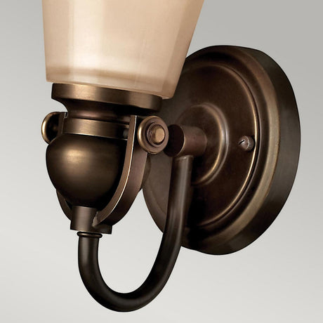 Close-up of the Mayflower 1 Light Wall Light - Bronze, showcasing a vintage-style bronze finish. The arm gracefully curves downwards to connect with a circular wall plate, supporting an amber frosted glass shade that radiates timeless charm.