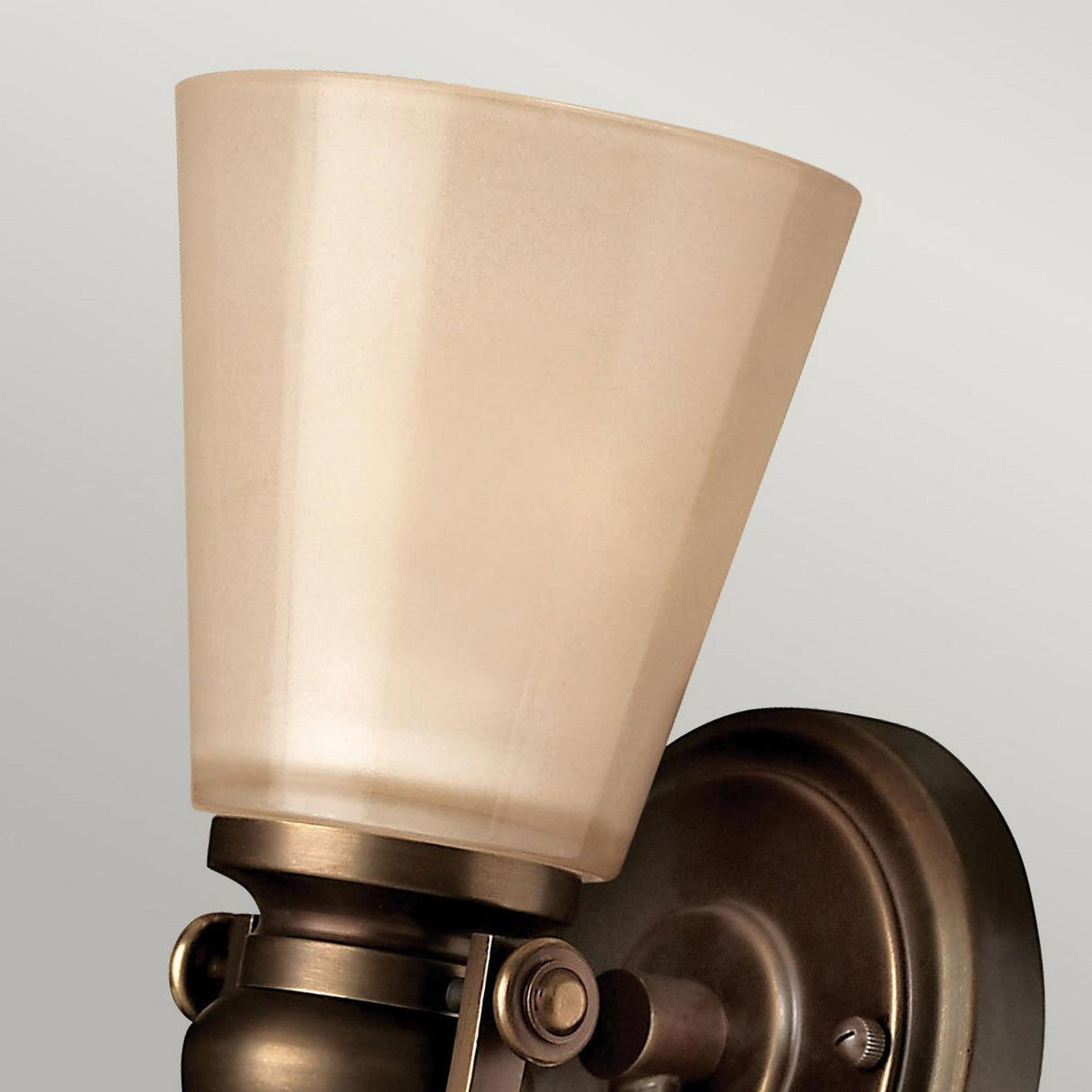 Close-up of the Mayflower 1 Light Wall Light - Bronze, showcasing its frosted glass shade and refined brushed bronze finish. The sconce features a round base and emits a warm, gentle glow against a light grey backdrop, echoing the inviting hue of amber glass.