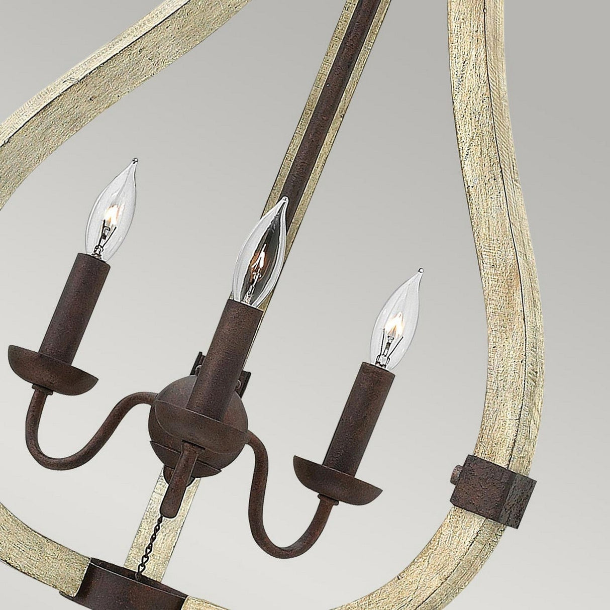 Introducing the Middlefield 3 Light Chandelier in Iron Rust, this piece showcases a rustic chic design with a wooden frame and metal candle-like bulb holders. Its teardrop-shaped bulbs enhance the elegant curved lines, all beautifully highlighted against a light gray background.
