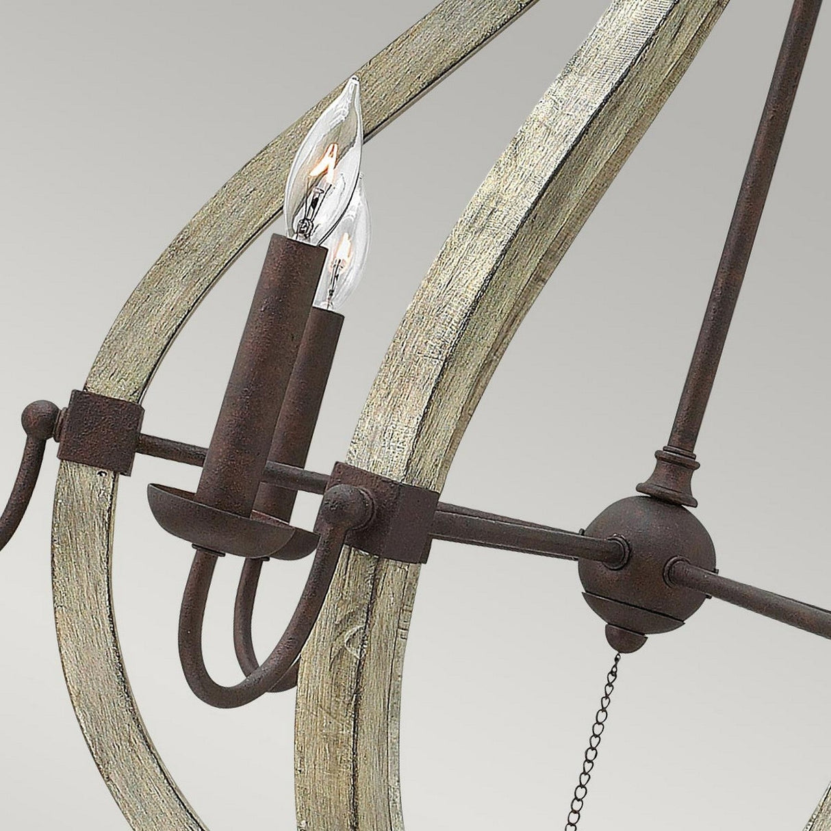 A close-up of the Middlefield 6 Light Chandelier - Iron Rust reveals a candle-shaped light bulb encased within a framework of distressed wood and metal. The iron rust finish provides an antique touch against the plain grey background.