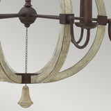 Close-up of the Middlefield 6 Light Chandelier, showcasing its distressed wood and charming iron rust finish. A small bell-like ornament hangs from the center, enhancing its antique allure against a plain, light gray background.