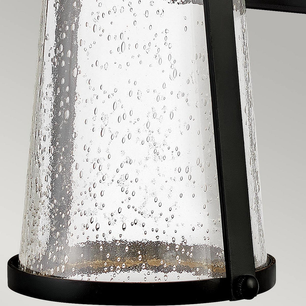 The Miles 1 Light Wall Lantern - Black features a cylindrical glass lamp with a bubbled texture, encased in black metal, embodying a minimalist yet nautical design. Its energy-efficient lighting softly reflects off the scattered bubbles, casting an elegant and decorative pattern.