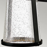 The Miles 1 Light Wall Lantern - Black features a cylindrical glass lamp with a bubbled texture, encased in black metal, embodying a minimalist yet nautical design. Its energy-efficient lighting softly reflects off the scattered bubbles, casting an elegant and decorative pattern.