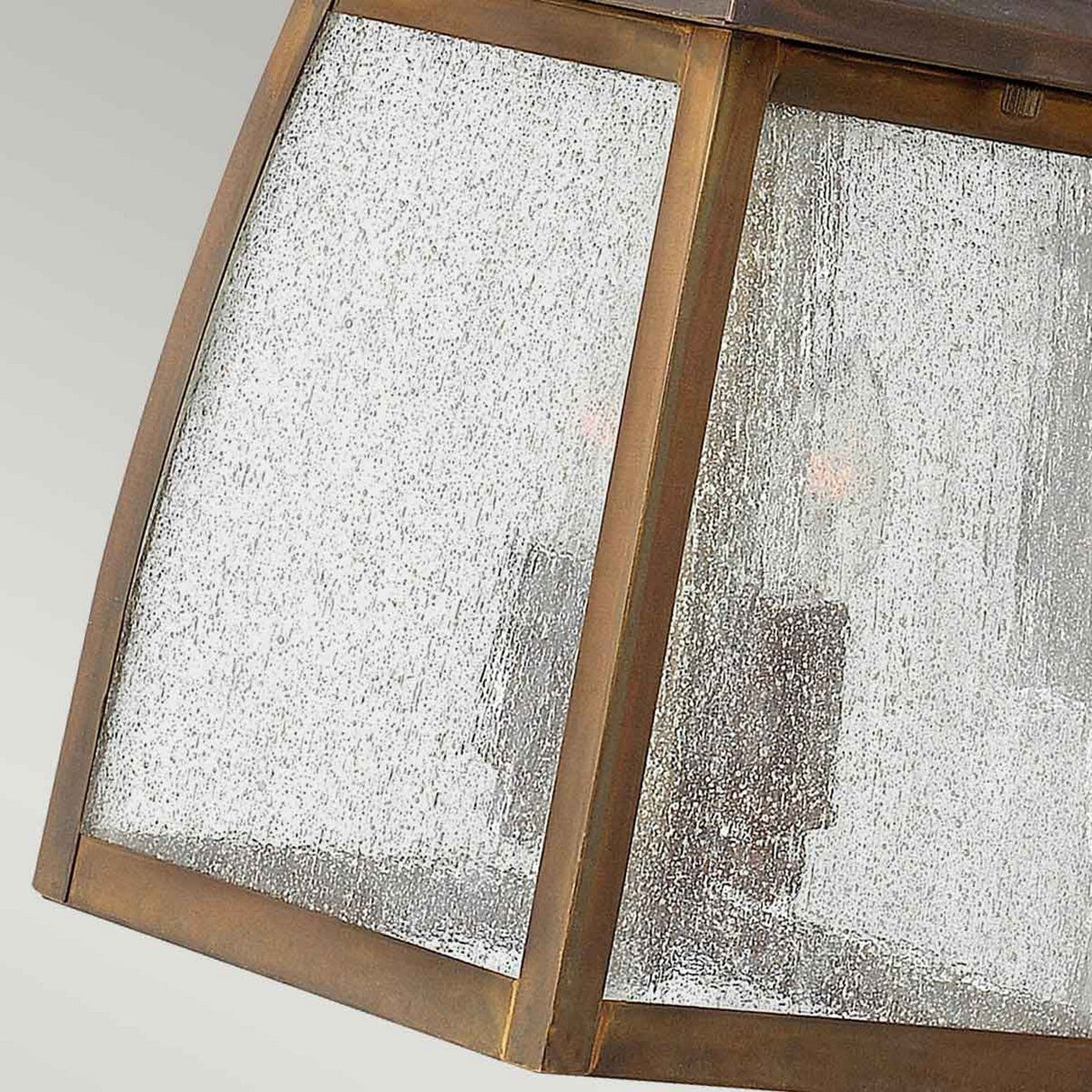 Close-up of the Montauk 2 Light Small Wall Lantern, featuring a solid brass frame and four textured glass panels. The glass, in a Sienna finish, has a speckled appearance that partially obscures the view of the two light bulbs inside. The background is a soft gradient.