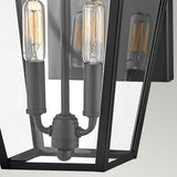 Close-up of the Palma Outdoor 2 Light Medium Wall Lantern in black, with clear glass panels displaying two vintage-style Edison bulbs. Its geometric trapezoidal frame artfully blends modern and classic elements, making it perfect as an outdoor wall lantern against a neutral gray backdrop.