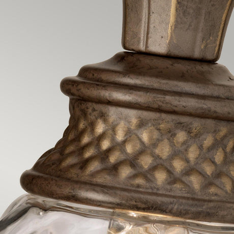 A detailed view of the Plantation Outdoor 1 Light Wall Light in a weather-resistant, antique bronze finish. The design showcases carved diamond patterns with a clear glass section below, set against a gray background.