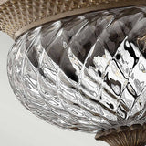 Close-up of a decorative, clear optic glass lampshade featuring a ribbed, swirling pattern. The top edge of the lamp has a textured design that complements the ornate bronze finish of the Plantation 2 Light Small Flush. The glass reflects light beautifully, producing intricate patterns and highlights.