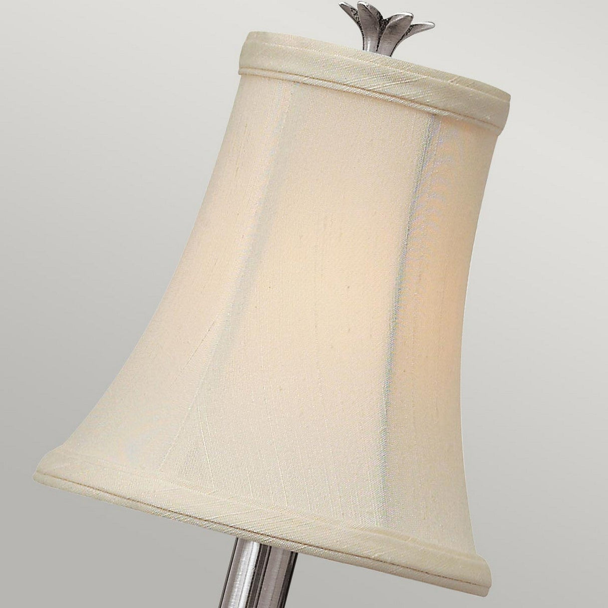 A close-up image showcases the Plantation 1 Light Wall Light with a cream-colored lampshade and a polished antique nickel stand. The slightly conical shade widens toward the top, adorned with a small decorative finial, against a neutral gray background that highlights its elegant metallic design.