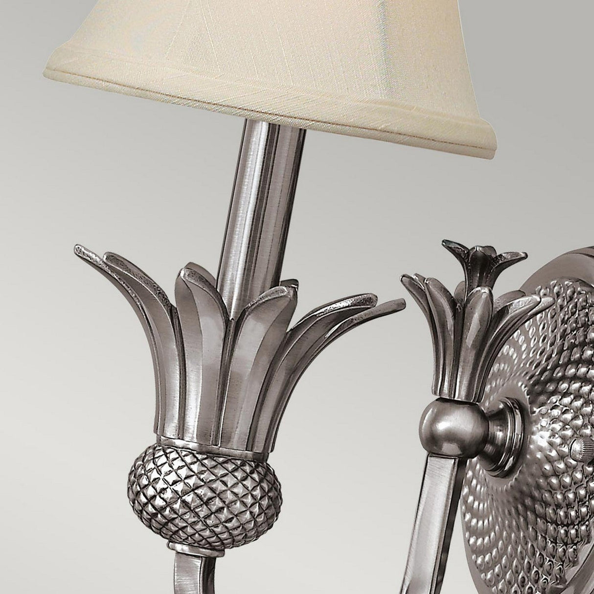 A detailed view of the Plantation 1 Light Wall Light in polished antique nickel, showcasing its elaborate flower-like design and textured base. The light is complemented by a light beige lampshade and placed against a neutral backdrop.