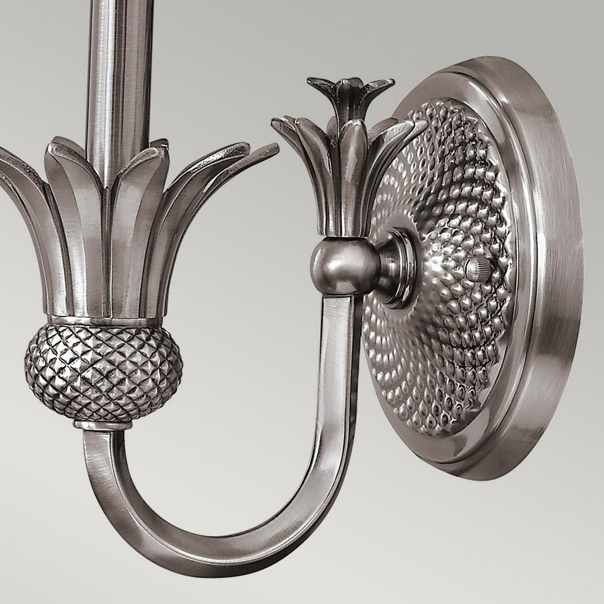 Close-up of the Plantation 1 Light Wall Light in polished antique nickel. The design showcases intricately patterned details that resemble leaves, along with a textured base, imparting an elegant, vintage appearance. Its smooth, polished finish accentuates the subtle reflections shimmering across the clear optic glass shade.