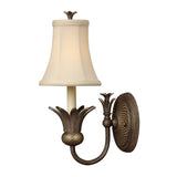 The Plantation 1 Light Wall Light - Bronze is an elegant wall-mounted fixture with a vintage-style design. It features a bronze finish, a flared fabric shade in white, and a metallic base adorned with ornate floral details. The light has a round, textured backplate and is shown turned off against a plain white background.