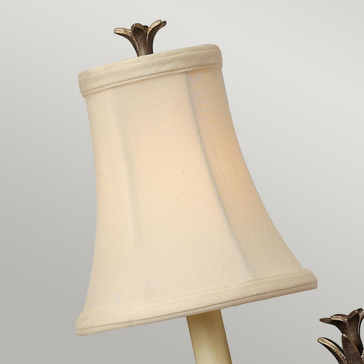 A close-up of a cream-colored lampshade features a stylized flower-shaped top finial, with the slightly flared design echoing the optic glass style seen in the Plantation 1 Light Wall Light - Bronze. The lampshade stands out beautifully against a plain background adorned with an elegant bronze finish.