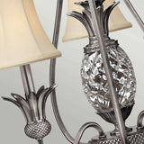 Close-up of the Plantation 3 Light Chandelier, featuring decorative metal work with pineapple motifs and beige fabric lampshades. The intricate design includes textured leaves and clear optic glass details, set against a soft gray background, capturing the elegance of the Plantation collection in its polished antique nickel finish.