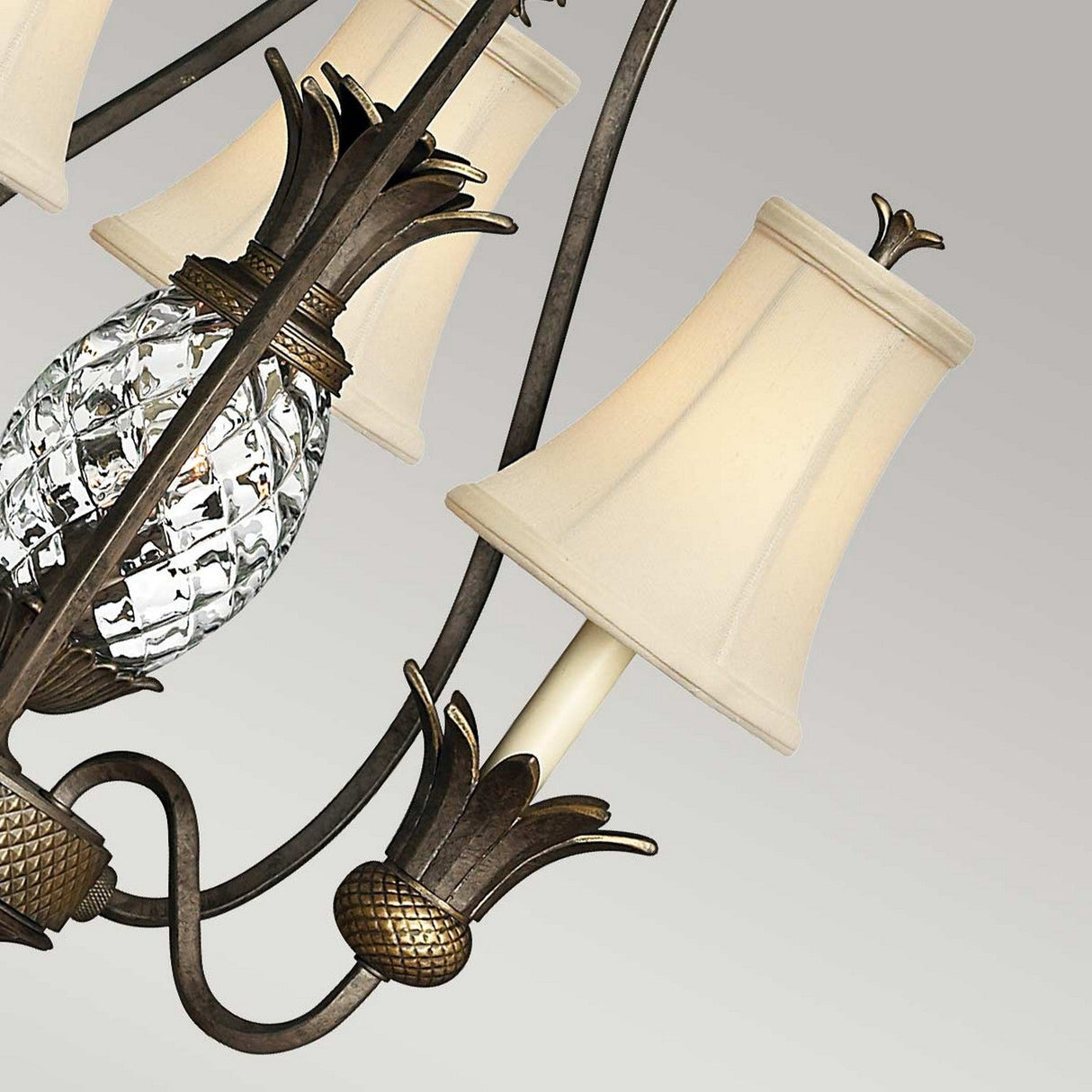 A close-up of the Plantation 3 Light Chandelier - Bronze reveals intricate crystal pineapple-shaped details and cream-colored fabric lampshades adorning a metallic frame with a Pearl Bronze finish. This chandelier highlights a harmonious blend of classic and elegant design elements against a neutral background.