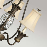 A close-up of the Plantation 3 Light Chandelier - Bronze reveals intricate crystal pineapple-shaped details and cream-colored fabric lampshades adorning a metallic frame with a Pearl Bronze finish. This chandelier highlights a harmonious blend of classic and elegant design elements against a neutral background.