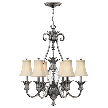 The Plantation 7 Light Chandelier in Polished Antique Nickel showcases six arms adorned with beige fabric lampshades and a central clear optic glass design shaped like a pineapple. Its detailed metalwork is elegantly suspended by a chain, making it perfect for sophisticated interior settings.