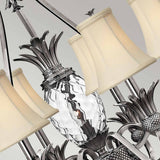 The Plantation 7 Light Chandelier - Polished Antique Nickel features a close-up view highlighting pineapple-shaped clear optic glass elements and multiple cream-colored lampshades set against a neutral background. Its intricate metal frame, crafted in polished antique nickel, exudes timeless elegance.