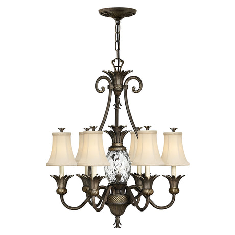 The Plantation 7 Light Chandelier in Pearl Bronze showcases intricate metalwork with seven arms, each adorned with a lamp featuring a white shade. It highlights a central pineapple-shaped clear optic glass detail, hanging gracefully from a chain.