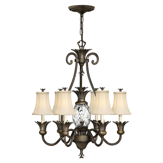 The Plantation 7 Light Chandelier in Pearl Bronze showcases intricate metalwork with seven arms, each adorned with a lamp featuring a white shade. It highlights a central pineapple-shaped clear optic glass detail, hanging gracefully from a chain.