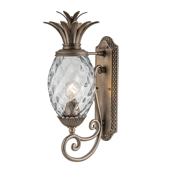 The Plantation Outdoor 1 Light Medium Wall Lantern features a decorative pineapple-shaped design and is wall-mounted. It has a burnished bronze finish, complemented by pineapple optic glass lamp housing and intricate detailing on the metal bracket and arm.