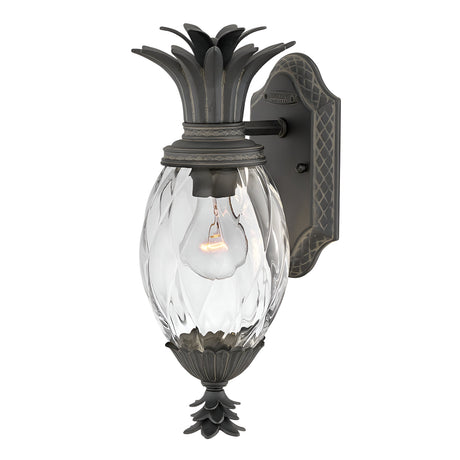 Introducing the Plantation Outdoor 1 Light Small Wall Lantern - Black, featuring a charming pineapple-shaped glass body with a clear, textured finish and dark metal accents. Its stylish black finish enhances its allure, allowing the visible bulb inside to cast an inviting glow. The backplate is adorned with a decorative pattern for added elegance.