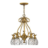 The Plantation 5 Light Chandelier - Burnished Brass is a vintage-style chandelier that showcases a burnished brass frame and five intricate crystal lampshades. Its design features scrolling metalwork adorned with decorative elements, including glass accents shaped like pineapples, and it is elegantly suspended by a chain.