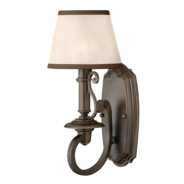 The Plymouth 1 Light Wall Light in bronze exudes vintage charm with its scrollwork design and oval base. Featuring a cream-colored, slightly tapered lampshade with dark brown trim, it embodies classic elegance, adding an exquisite touch to your interior decor.