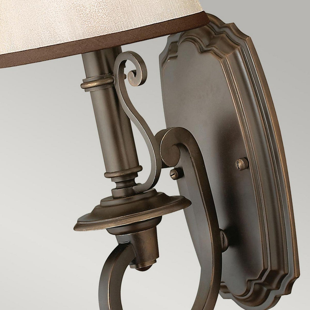 Close-up of the Plymouth 1 Light Wall Light in bronze, featuring intricate metalwork and a beige lampshade against a gray background. The design exudes classic elegance with its decorative scrollwork and scalloped backplate, adding vintage charm to any space.